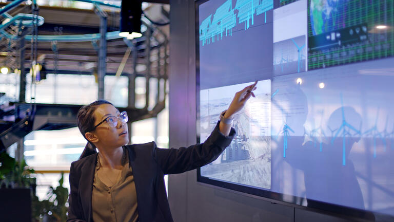 Stock photograph of a young Asian woman conducting a seminar / lecture with the aid of a large screen. The screen is displaying data & designs concerning low carbon electricity production with solar panels & wind turbines. These are juxtaposed with an image of conventional fossil fuel oil production.