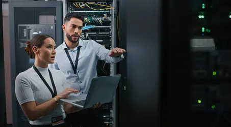 Two professionals working in a server room, looking at a laptop