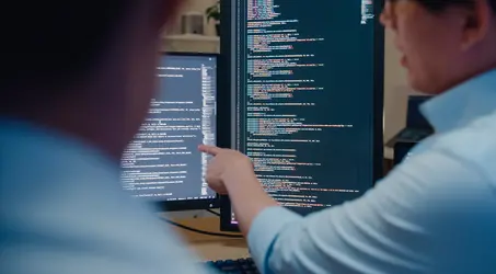 Two professionals reviewing lines of code on a computer monitor
