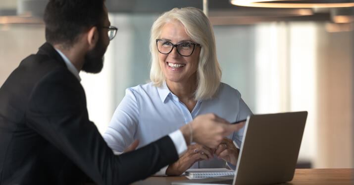 Businessman consulting mature businesswoman client about loan, insurance or investment, using laptop, pointing at screen, young man helping senior colleague with software, employee presenting project