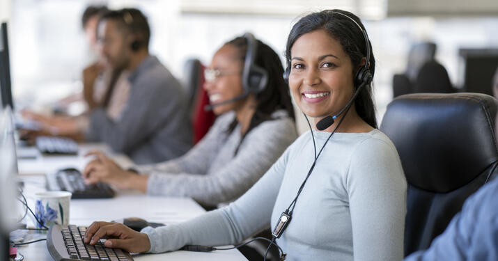 Portrait of smiling businesswoman wearing headset while using computer. Confident female operator is working with colleagues at desk. They are in call center.